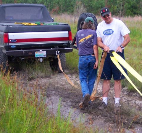 1988 Ford Bronco - Two people attach tow straps to a lifted Ford Bronco on a dirt path.