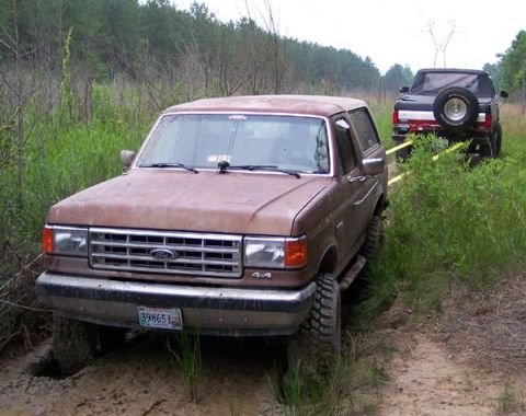 1988 Ford Bronco - Two Ford Broncos are on a muddy trail, with one vehicle stuck and being towed by the other.