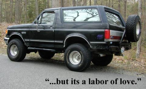 1988 Ford Bronco - This black Ford Bronco is parked on a paved road with trees in the background.