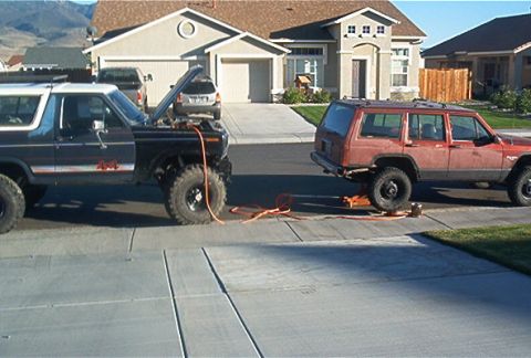 1986 Ford Bronco - Two SUVs, a Ford Bronco and a Jeep Cherokee, are parked side-by-side in a driveway with work being done on them.