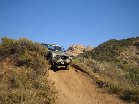 1990 Ford Bronco - Green Ford SUV climbing a steep dirt trail with rock formations behind it under a clear blue desert sky.