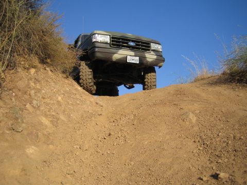 1990 Ford Bronco - Dramatic low-angle shot of a green lifted Ford truck cresting a dirt mound off-road against a blue sky.
