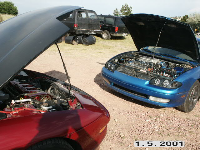 Red and blue 1990s coupes with hoods open on a dirt lot, engine bays exposed