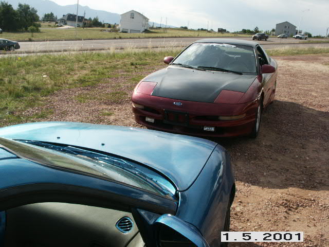 Red Ford Probe with flat black hood on gravel, seen past a teal car's hood