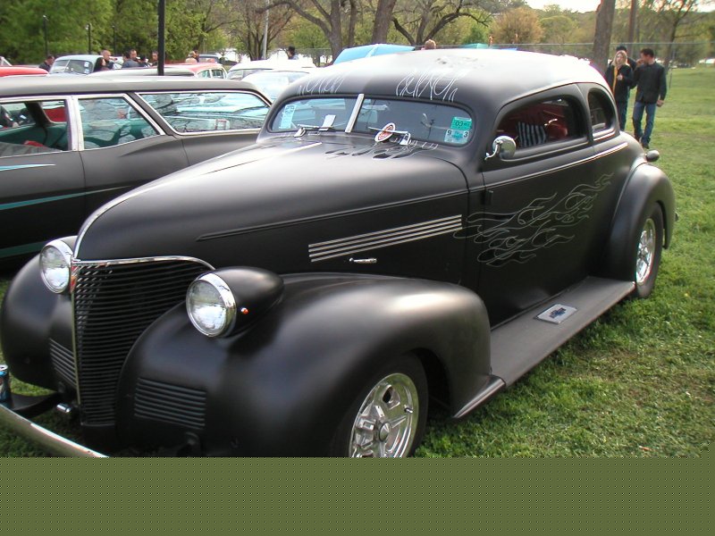 Matte black custom hot rod coupe with silver flame graphics and pinstriping on the windshield. Another car is visible to the left