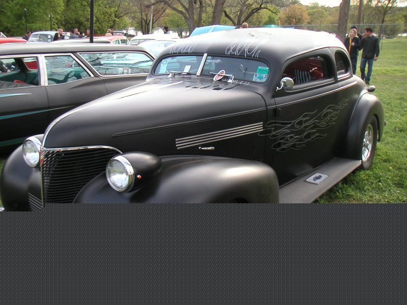 Black hot rod coupe with flame decals and pinstriping on the windshield. Another car is parked next to it