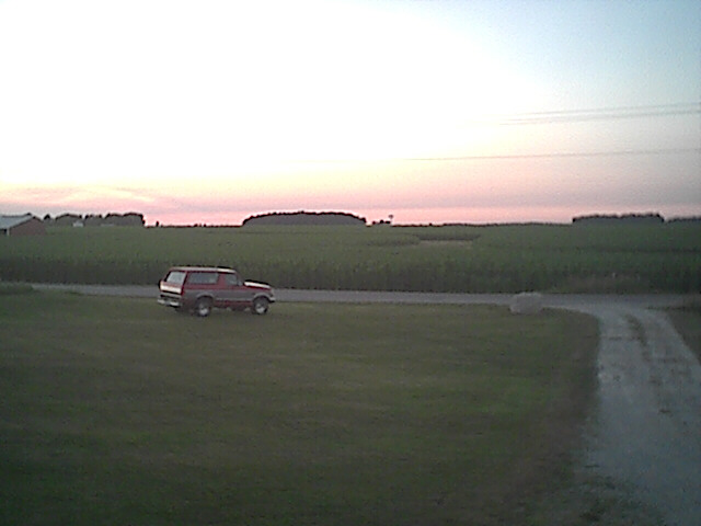 Red 1994 Ford Bronco side view driving on a dirt road with fields in background