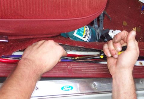 1985 Ford LTD Crown Victoria - Hands work on wiring beneath the red carpet of a vehicle's interior, revealing a Ford logo on the door sill.