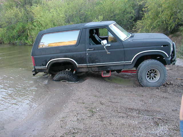 Black Ford Bronco with one rear wheel submerged in water, stuck on a muddy bank