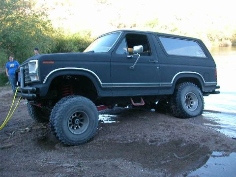 1984 Ford Bronco - This black Ford Bronco, likely from the early 1980s, is positioned on a muddy bank near the water.