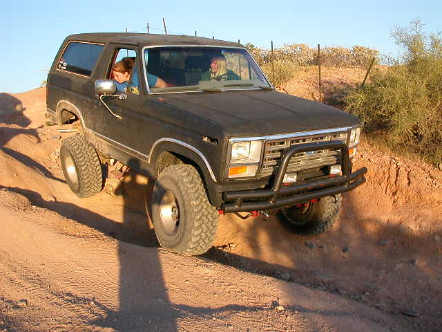 1984 Ford Bronco driving on a dirt trail with two people inside
