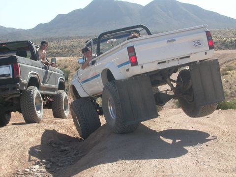 1984 Ford Bronco - Two modified trucks navigate a challenging off-road course in a desert landscape.