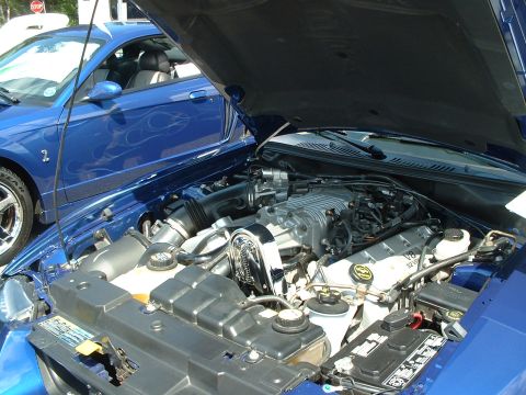 1999 Ford Crown Victoria - The engine bay of a blue Ford Mustang, a prominent supercharger and a second Mustang with flame decals in the background.