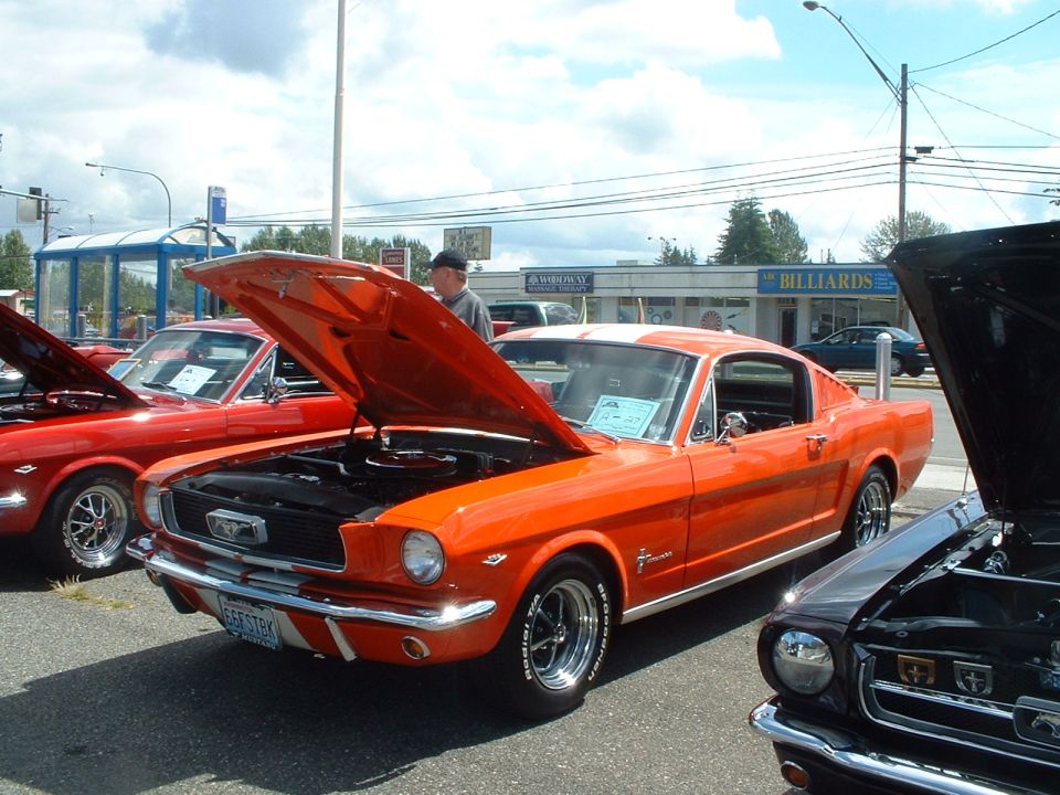 Orange 1965 Ford Mustang fastback with hood open, parked next to other classic cars