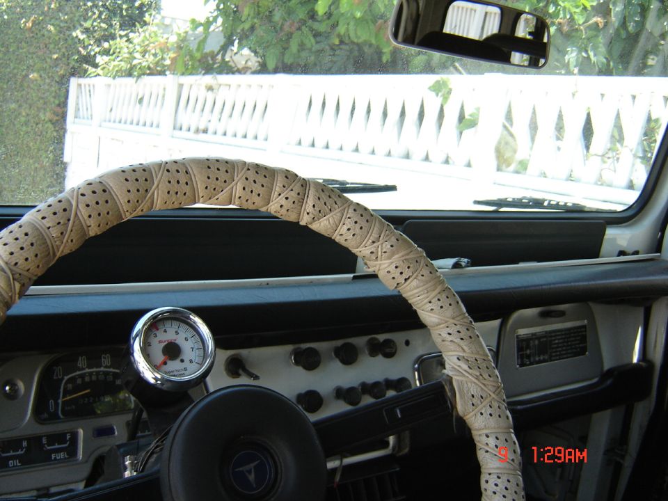 1977 Toyota Land Cruiser interior with a beige steering wheel cover, dashboard gauges, and a white fence visible through the windshield