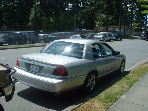 1999 Ford Crown Victoria - Silver sedan parked on street with other vehicles in the background.