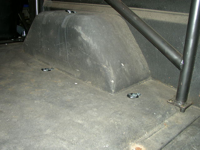 Interior cargo area of a vehicle, showing the wheel well and several tie-down rings on the floor. A roll cage is visible