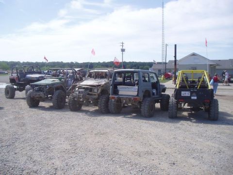 1992 Ford Bronco - A collection of heavily modified off-road vehicles are lined up in a gravel lot under a partly cloudy sky.