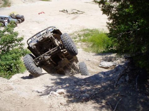 1992 Ford Bronco - An off-road vehicle is stuck on its side while navigating a challenging dirt terrain, with other vehicles visible in the background.
