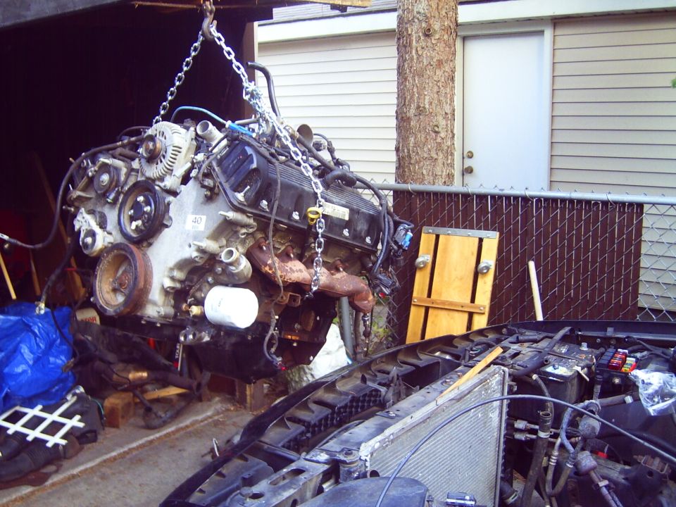Engine suspended by chains, with a car's open hood and radiator visible below