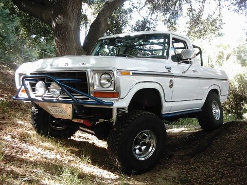 1978 Ford Bronco - This lifted 1978 Ford Bronco features a blue front bumper with auxiliary lights and off-road tires, parked on a dirt path.