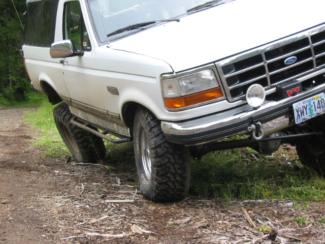 White lifted Ford Bronco with large mud tires parked on a dirt road, front three-quarter view.