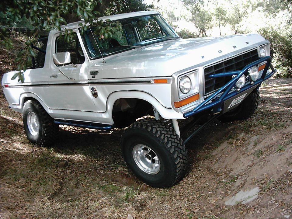 Silver 1978 Ford Bronco with blue off-road bumper and large tires, parked on dirt