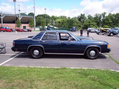 2001 Ford Crown Victoria - A dark blue sedan is parked in a lot near a stadium, with other vehicles and people in the background.