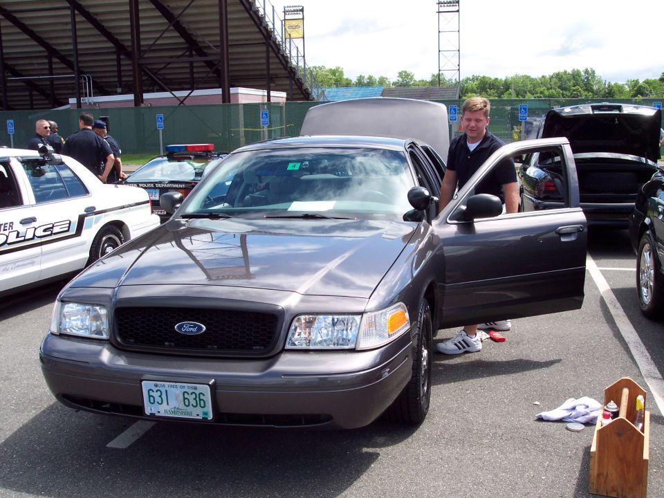 Gray Ford Crown Victoria with driver's door open, parked next to a white police car. Guy in black shirt stands by the open door