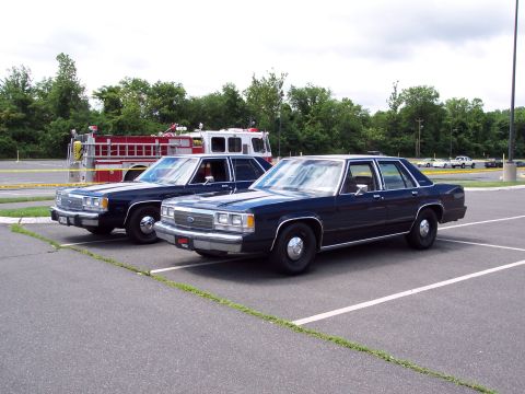 2001 Ford Crown Victoria - Two dark blue police sedans are parked in a lot, with a fire truck visible in the background.