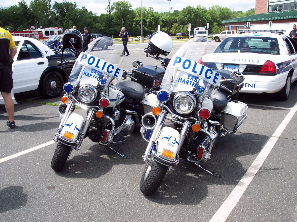 Two police motorcycles parked in a lot, with a police car in the background. The motorcycles have 'POLICE' and 'EAST HARTFORD' on their windshields