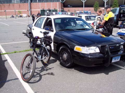 2001 Ford Crown Victoria - A police car and a bicycle are parked in a lot, with officers standing around.