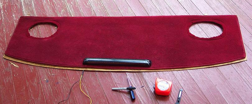 Red carpet piece with two circular cutouts and a black vent strip, lying on a wooden floor next to tools