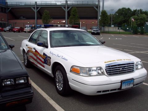 2001 Ford Crown Victoria - A white 2001 Ford Crown Victoria police car, marked with an American flag decal and "Building on a Proud Tradition" text, is parked next to…