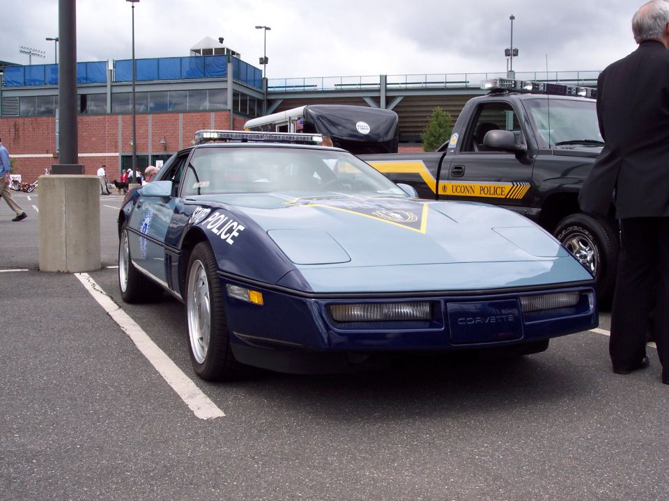 Blue and light blue police Corvette parked next to a black pickup truck with yellow stripes and 'UCONN POLICE' text