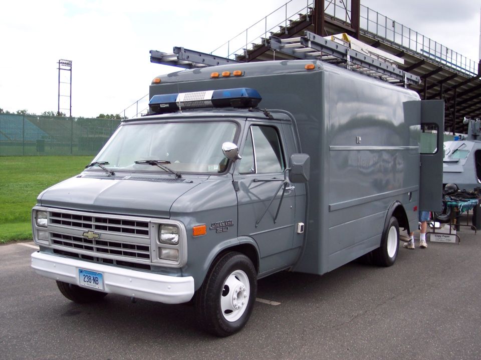 Gray utility van with a boxy rear section, ladder rack on top, and blue lights on the roof. License plate reads 238 NB