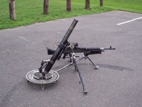 2001 Ford Crown Victoria - A mortar and machine gun system is deployed on a tripod on an asphalt surface, with grass and trees in the background.