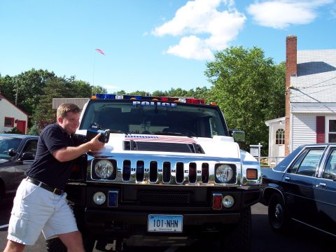 2001 Ford Crown Victoria - A man aims a radar gun at a Hummer H2 SUV marked as a police vehicle, with another police car visible to the side.
