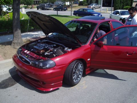 1993 Ford Taurus - A red sedan is parked on a street with its hood and driver's door open, revealing the engine.
