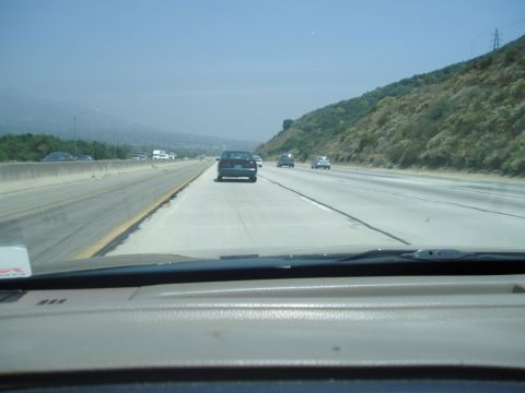 1993 Ford Taurus - A dark sedan travels down a multi-lane highway, with other vehicles in view and a tree-covered hill to the right.