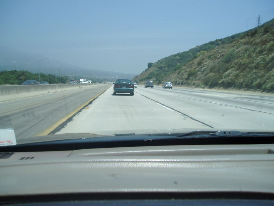 Dark sedan seen from behind on a highway, with other cars and a hilly landscape