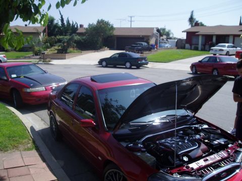 1993 Ford Taurus - A red 1993 Ford Taurus is parked with its hood open, revealing the engine bay and a strut bar, while other vehicles are visible on the stre…