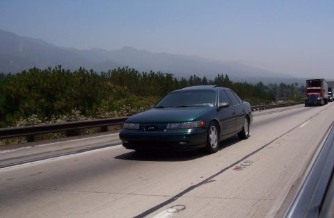 1993 Ford Taurus - A green sedan is captured from a side angle on a highway with mountains visible in the distance.
