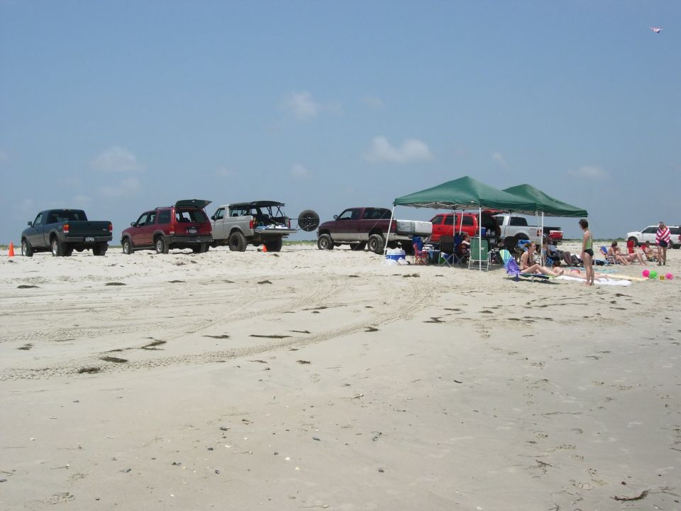 Row of 4x4 trucks and SUVs parked on a sandy beach beside green pop-up canopies and beachgoers