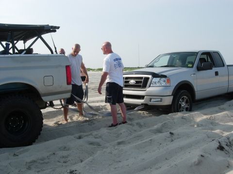1995 Ford Bronco - Two trucks are stuck in the sand on a beach, with people attempting to pull one out.