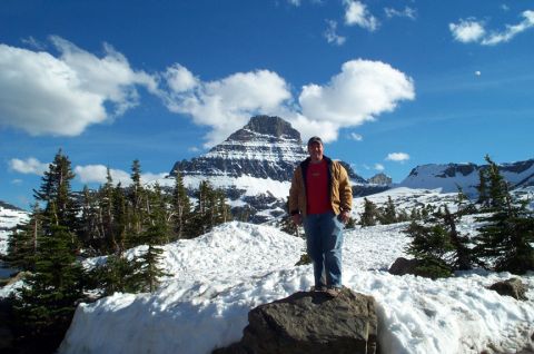 1996 Ford Bronco - A man poses for a photo in a scenic mountain landscape with snow-covered peaks and trees under a blue sky with clouds.
