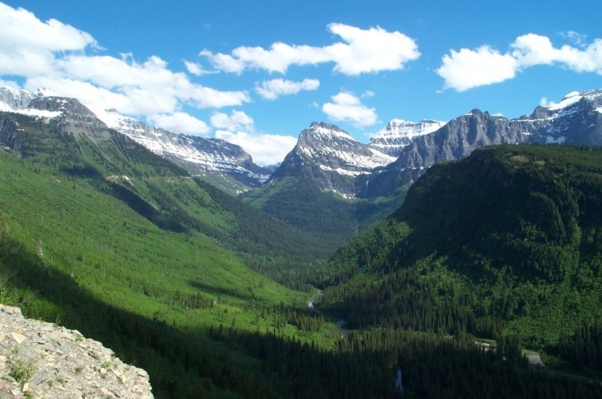 Mountain valley with snow-capped peaks, green forested slopes, and a winding river under a blue sky with clouds. Watermark in lower right