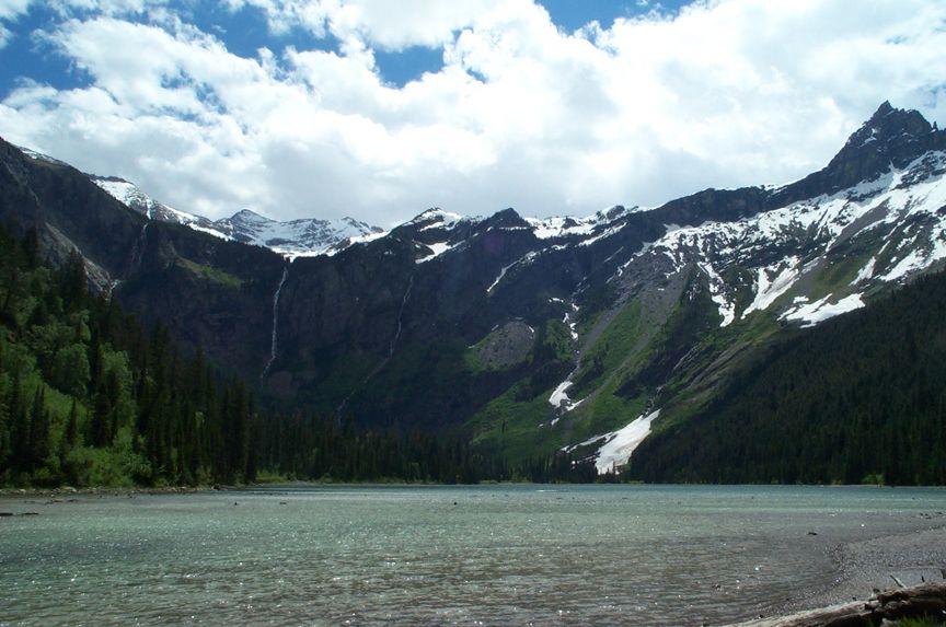 Mountain range with snow-capped peaks, evergreen forest, and a clear lake under a cloudy sky