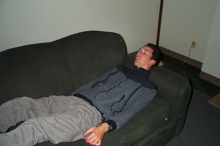guy passed out on a dark green couch, wearing a gray jacket and tan pants. He has a watch on his left wrist