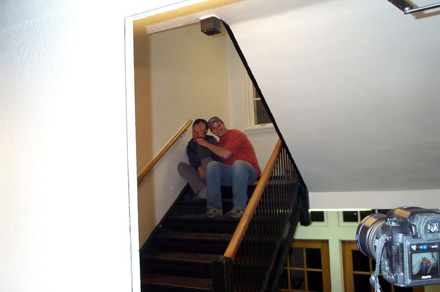 Two people sit on a wooden staircase, one with an arm around the other. A camera is in the foreground